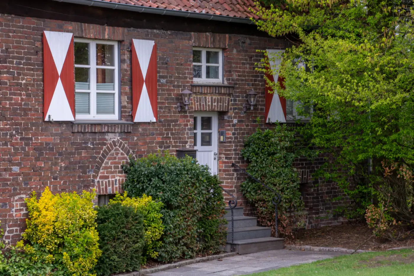 Backsteinhaus mit rot-weißen Fensterläden, weißer Haustür und Treppe zwischen Sträuchern (Zusamaltheim, Landkreis Dillingen an der Donau)